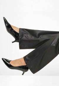 Escarpins à talons hauts en cuir verni noir avec des bouts pointus, associés à un pantalon noir à texture lisse et évasée, mettant en valeur un design épuré.