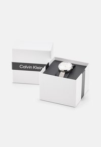Reloj con correa de malla plateada y esfera blanca, colocado en una caja blanca abierta que presenta una banda negra con "Calvin Klein" impresa en ella.