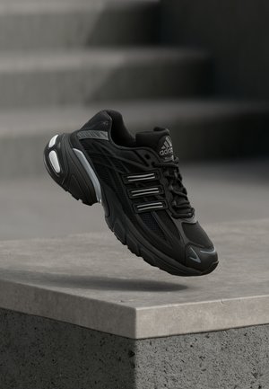 Chaussure de course Adidas noire avec des détails réfléchissants flottant au-dessus d'une marche en béton sur un fond gris flou.