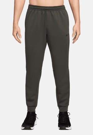 Mann trägt dunkelgraue Nike-Sweathosen und schwarze Sportschuhe mit weißen Sohlen, steht vor einem schlichten hellen Hintergrund.