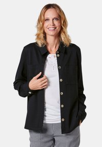 Zwarte button-up shirt van gladde stof, met een kraag, twee borstzakken en gouden knopen. Gekocht over een witte top met kantaccenten.