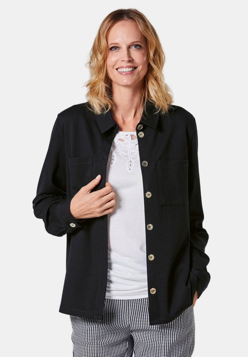 Zwarte button-up shirt van gladde stof, met een kraag, twee borstzakken en gouden knopen. Gekocht over een witte top met kantaccenten.