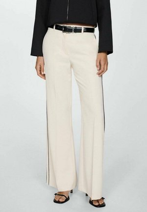 Mujer con pantalones anchos color crema con franjas negras laterales, cinturón negro, chaqueta negra corta y tacones abiertos negros, de pie frente a un fondo liso.