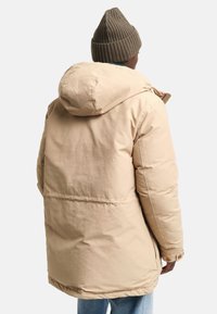 Beżowa kurtka puchowa z kapturem, wykonana z teksturowanej tkaniny. Charakteryzuje się prostym fasonem, widocznymi szwami i bocznymi kieszeniami. Na głowie założona jest prążkowana khaki beanie.