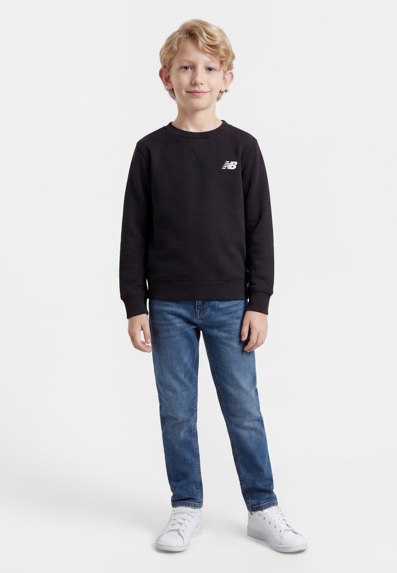 Ξανθός αγόρι που στέκεται, φοράει μαύρο sweatshirt New Balance, μπλε τζιν και λευκά αθλητικά παπούτσια, μπροστά σε λευκό, απλό φόντο.