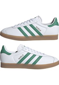 Weiße Leder-Sneaker mit grünen Akzenten und drei Streifen-Design. Mit einer Gummisohle und dem "GAZELLE"-Logo an der Seite. Klassische Form.
