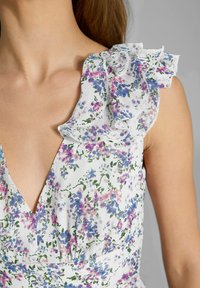 Nahaufnahme einer Frau, die ein weißes Blumenkleid mit lila und rosa Blumen trägt, mit rüschendem Stoff auf einer Schulter und einem tiefen V-Ausschnitt.