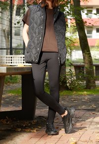Černá prošívaná vesta přes hnědou košili, doplněná černými legínami a strukturovanými černými teniskami, prezentována v venkovním prostředí s listy.