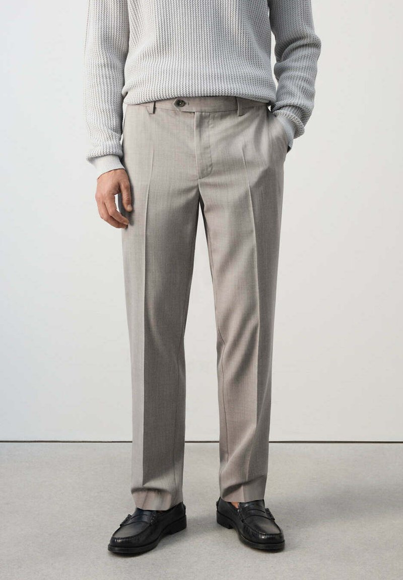 Homme portant un pantalon de costume gris clair, des mocassins noirs et un pull tricoté gris clair, debout avec une main dans la poche.