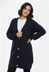 Kobieta o krótkich brązowych włosach, ubrana w granatowy, długi, dziergany kardigan z dużymi guzikami na białej koszuli, uśmiechająca się, z jedną ręką na biodrze.