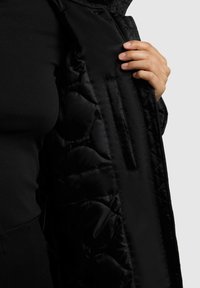 Melnā pildītā jakā ar kvadrātveida faktūru, kas aprīkota ar sānu rāvējslēdzēju kabatu un gludu, spīdīgu auduma tekstūru. Tuvplāns skats uz iekšpusi.