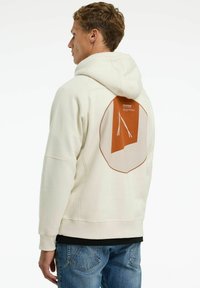 Crème hoodie met een gestructureerde oranje patch op de achterkant, voorzien van een geometrisch ontwerp en trektouwtjes. Ribgebreide manchetten en onderkant.