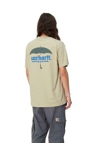 Camiseta de algodón beige con un gran gráfico de paraguas azul y el texto "carhartt. cubriendo tus necesidades" en la parte posterior. Mangas cortas, ajuste relajado.