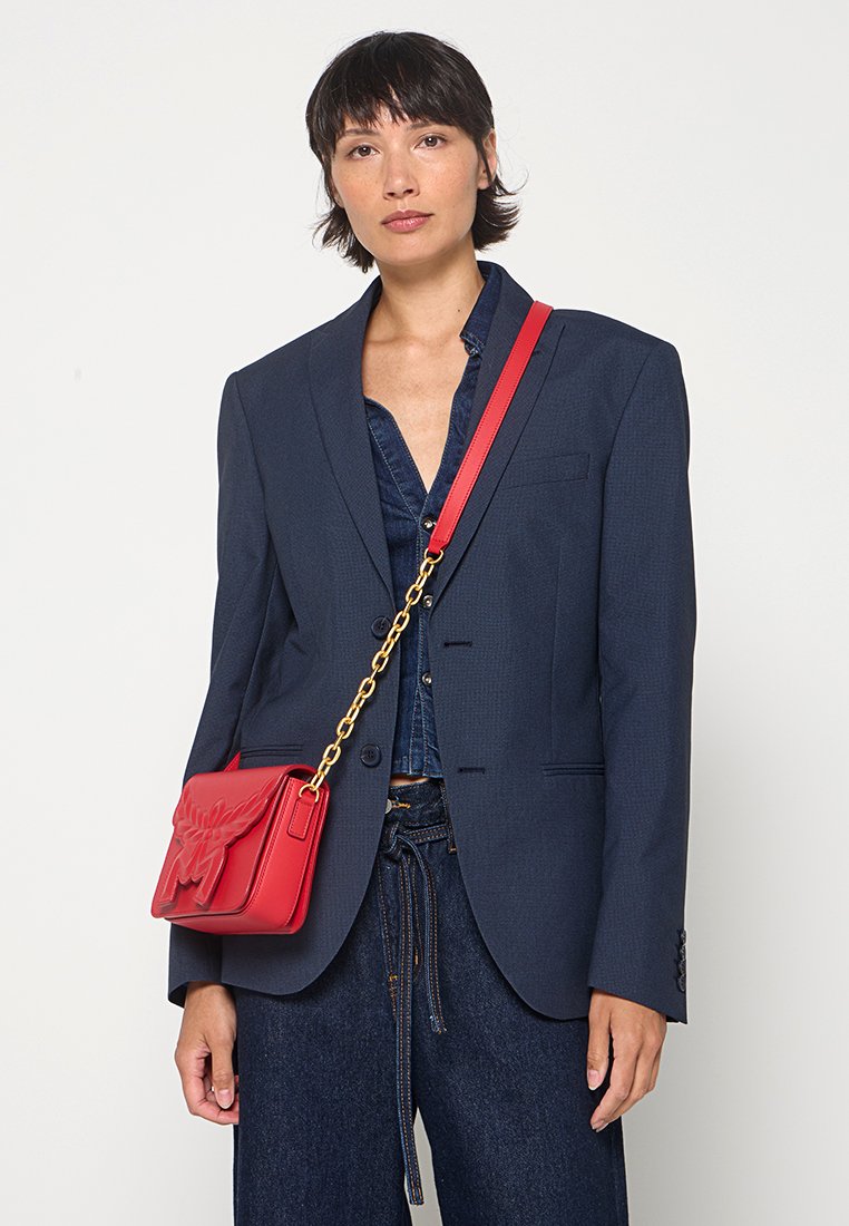 Rote Leder-Schultertasche mit geprägtem Blumenmuster und goldener Kettenriemen, getragen zu einem marineblauen Blazer und einem Jeanshemd, vor einem weißen Hintergrund.