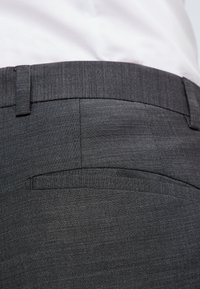 Grå texturerade byxor med flat front, bälteshällor och detalj av bakficka. Tyget visar ett subtilt mönster. Sömsömmarna är synliga.