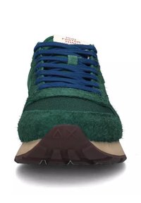 Sneaker verde con tomaia texturizzata e pelosa e pannelli in rete. Lacci blu, linguetta crema con testo rosso e suola in gomma marrone per una buona aderenza.