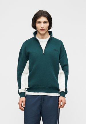 Mladić u tamnozelenoj i bijeloj majici s patentnim zatvaračem i tamnoplavim hlačama stoji ispred jednobojne svijetlosive pozadine.