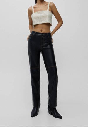 Woman wearing a white beaded cropped tank top, high-waisted black leather pants, and black pointed-toe boots, standing with hands behind back.