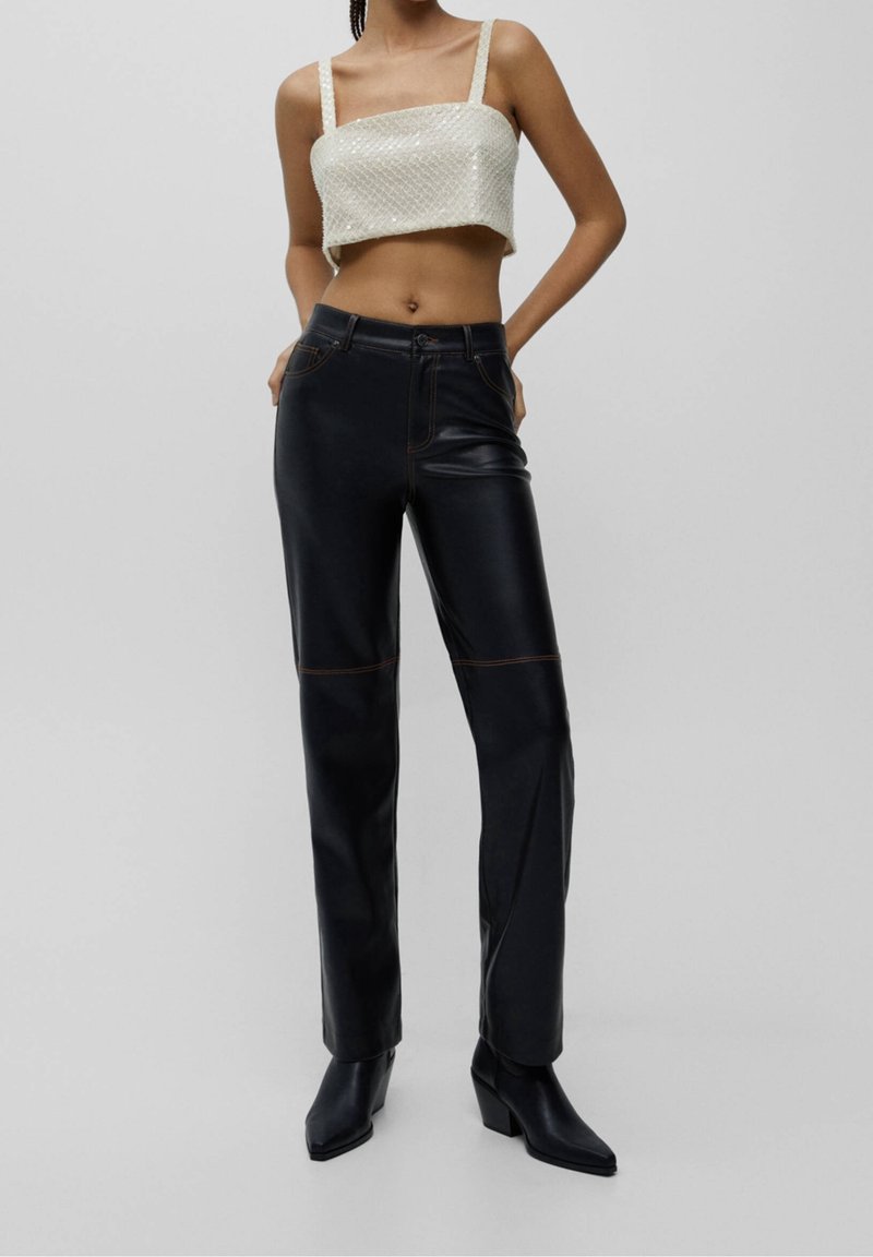 Femme portant un top court blanc orné de perles, un pantalon en cuir noir taille haute et des bottes noires à bout pointu, debout les mains dans le dos.