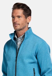 Lichtblauwe zip-up jas met een hoge kraag, voorzien van een textuurstof en een zwarte rits, gelaagd over een kraag van een bloempatronen shirt.