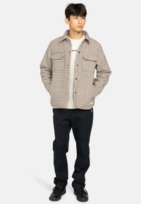 Rutet skjortejakke i beige og brune nuancer, der har to brystlommer, knaplukning foran og en afslappet pasform, parret med sorte bukser og sneakers.