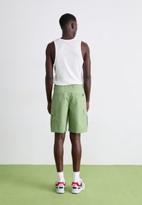 Uomo in piedi di fronte a una parete bianca, che indossa una canotta bianca, pantaloni cargo leggeri verdi, calzini bianchi e sneakers bianche e rosse su un pavimento verde.