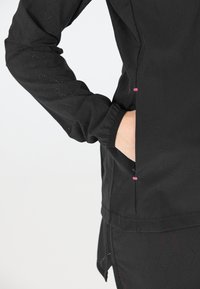 Svart idrottsjacka med perforerade ärmar och rosa detaljer. Har en sidoficka och elastiska ärmar för en skräddarsydd passform.