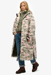 Imperméable camouflage avec motifs roses et gris, design long et capuche. Porté sur une veste matelassée verte et un jean bleu avec des bottes marron.