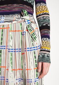 Pull multicolore avec un motif rayé, associé à une jupe plissée à lignes verticales et un nœud papillon dans des couleurs vives.