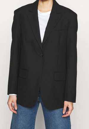 Person trägt einen schwarzen Blazer mit einem Knopf über einem weißen Hemd und blauen Jeans, steht vor einem einfarbigen hellen Hintergrund.