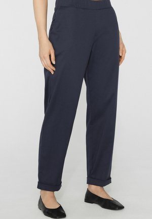 Frau trägt marineblaue, umgeschlagene Hose mit elastischem Bund und schwarze flache Schuhe, steht auf einem hellgrauen Boden.