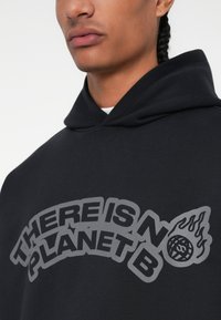 Μαύρο φούτερ από μαλακό ύφασμα, με μεγάλο γκρι κείμενο "ΔΕΝ ΥΠΑΡΧΕΙ ΣΧΕΔΙΟ Β" και γραφικό φλόγας. Χαλαρή εφαρμογή με κουκούλα που έχει κορδόνι.