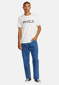 Hvid bomulds-t-shirt med sort "RVCA" logo, parret med blå denim jeans og sorte lavtop sneakers. Enkel design og afslappet pasform.