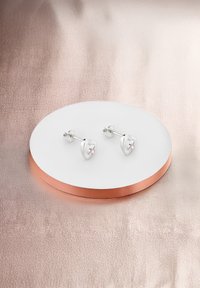 Silvriga hjärtformade studs med en rosa accentsten, utställda på en rund vit och kopparfärgad hållare mot en mjuk, texturerad bakgrund.