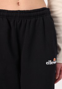 Melni kokvilnas sporta bikses ar elastīgu jostasvietu, uz kurām ir balts izšūts "ellesse" logotips un krāsains logotipa akcenti tuvu kabatai.