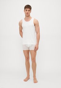 White tank top and sheer white boxer briefs on a standing male model, showing fabric texture and form-fitting design. Solid colors, no patterns.