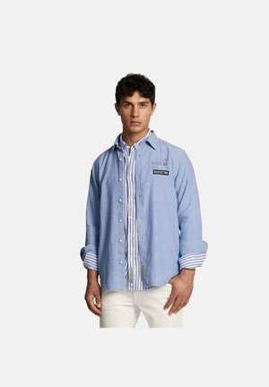 Mann trägt hellblaues Hemd mit Knöpfen über einem weiß-blau gestreiften Hemd, mit weißen Hosen, steht vor einem schlichten Hintergrund.