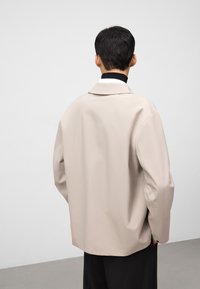 Beige oversized jas met een zachte textuur, met een brede kraag en knoopmanchetten, gedragen over een zwarte coltrui, gezien vanaf de achterkant.