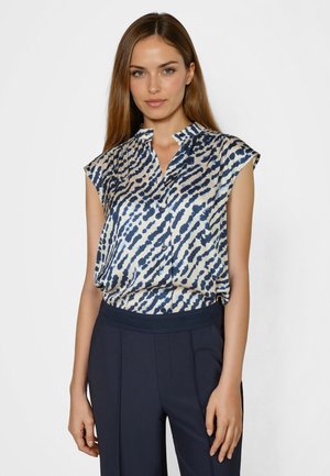 Woman wearing sleeveless, navy blue and white patterned button-up blouse with dark navy high-waist pants, standing against plain background.