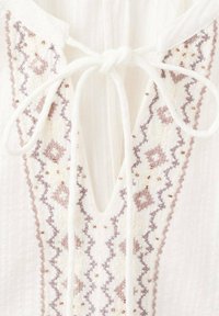 Blusa blanca con escote en V y bordados intrincados en marrón claro y malva, adornada con pequeñas cuentas y lazos en el cuello.