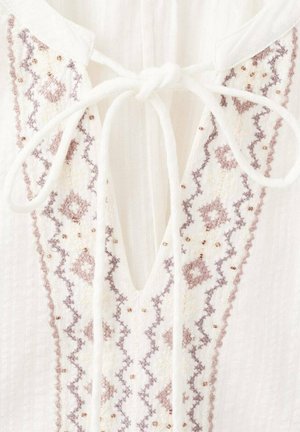 Weiße Bluse mit V-Ausschnitt, verziert mit filigraner Stickerei in Hellbraun und Mauve, akzentuiert mit kleinen Perlen und Bändchen am Halsausschnitt.