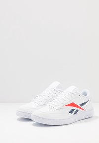 Vita låga sneakers med marinblå och röda geometriska sidopunkter samt vita snörningar på en helt vit yta och bakgrund.