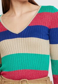 Dziany, wielokolorowy sweter z poziomymi paskami w kolorach czerwonym, niebieskim, zielonym i beżowym. Dekolt w serek z prążkowaną fakturą.