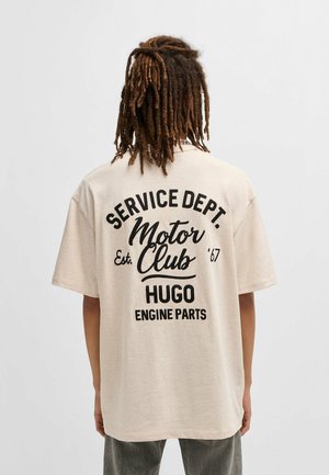 Beige oversized t-shirt featuring black text print on the back: "SERVICE DEPT. Motor Club Est. '67 HUGO ENGINE PARTS." Short sleeves, relaxed fit.