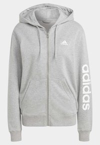 Šedá mikina s kapucí na zip vyrobená z textilu, s předním zipem, dvěma bočními kapsami a bílým nápisem "adidas" vytištěným na levém rukávu.