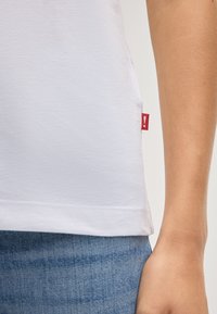 Weißes Baumwoll-T-Shirt mit abgerundetem Saum, versehen mit einem kleinen roten Etikett an der Seite. Getragen mit Denim, zeigt Stoffstruktur und Passform.