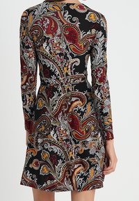 Vestido de manga larga con cintura ajustada y patrón colorido de paisley en rojo, mostaza y blanco sobre un fondo negro.