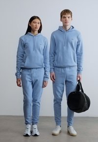 Ανοιχτό μπλε ταιριαστά hoodies και παντελόνια φούτερ από απαλό ύφασμα, με τσέπες μπροστά και κορδόνια· συνοδεύονται από μια μαύρη στρογγυλή τσάντα.