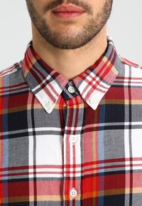 Camicia in cotone a quadri con colori rosso, nero, bianco e blu. Collo button-down e bottoni bianchi con un motivo tessuto testurizzato.