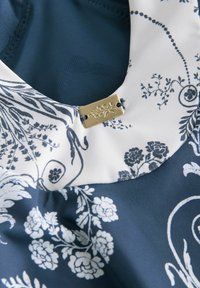 Kukallinen kangas tummansinisin ja valkoisin värein, jossa on pyöreä kaula-aukko ja kultainen akcentti kauluksen lähellä.